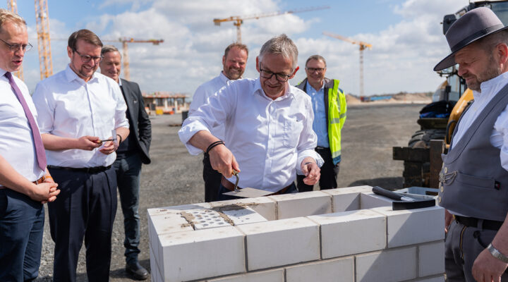Landrat Olaf Meinen setzt mit einer Kelle Mörtel auf die Steine eines gemauerten Fundaments, während die anderen zuschauen.