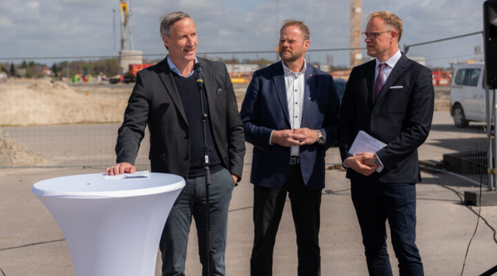 Dirk Balster, Geschäftsführer der Trägergesellschaft Kliniken Aurich-Emden-Norden mbH, Matthias Arends, Landtagsabgeordneter (MdL) und Wiard Siebels, Landtagsabgeordneter (MdL) in Anzügen stehen im Freien auf einer Baustelle; einer spricht an einem Mikrofon, während die anderen daneben stehen.