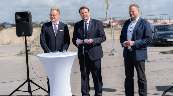 Landtagsabgeordneter Wiard Siebels, Oberbürgermeister Tim Kruithoff und Landtagsabgeordneter Matthias Arends in Anzügen stehen im Freien auf einer Baustelle; einer spricht an einem Mikrofon, während die anderen daneben stehen.