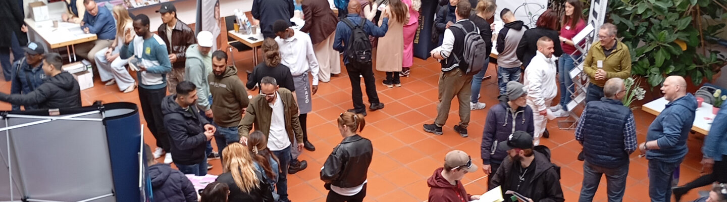 Viele Menschen besuchen eine Jobmesse in einem Innenraum, stehen an Informationsständen, sprechen miteinander und informieren sich über Angebote.