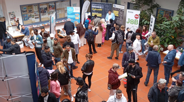 Viele Menschen besuchen eine Jobmesse in einem Innenraum, stehen an Informationsständen, sprechen miteinander und informieren sich über Angebote.