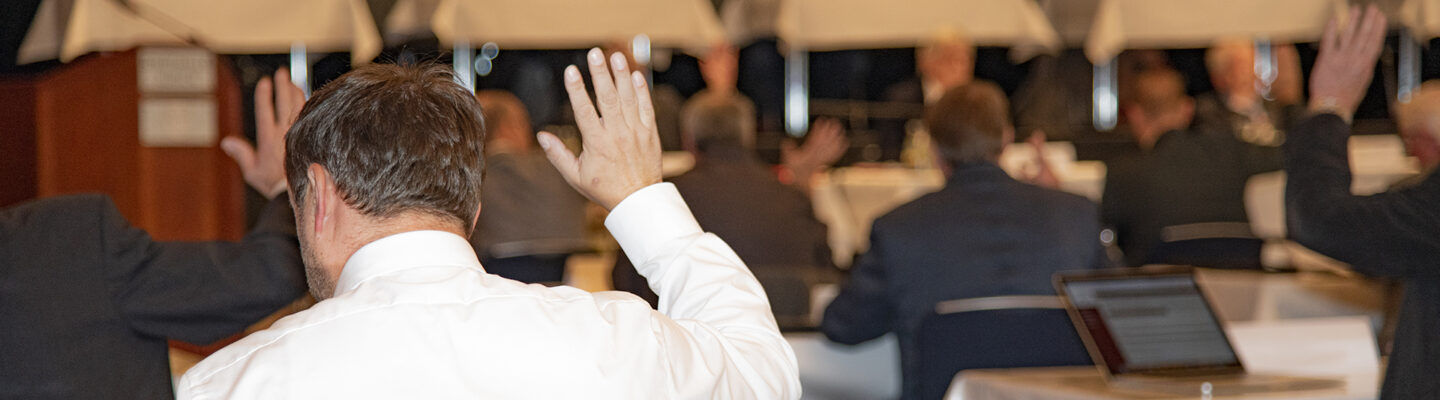 Mehrere Personen sitzen in einem Saal an Tischen und heben die Hand, während vorne auf einer Bühne ein Podium mit mehreren Rednern zu sehen ist.
