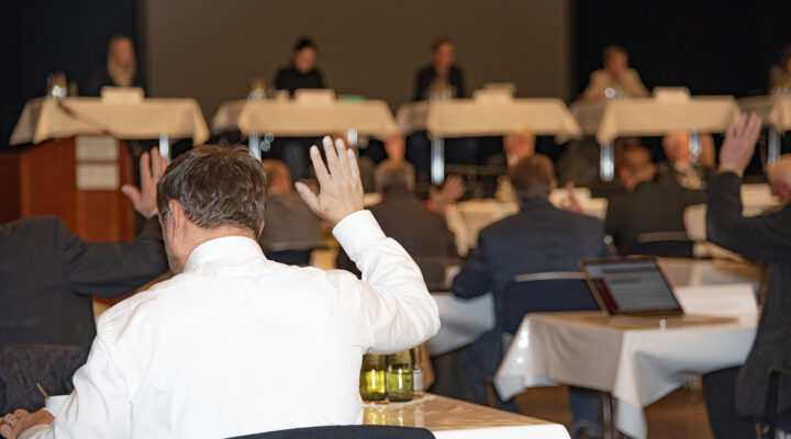 Mehrere Personen sitzen in einem Saal an Tischen und heben die Hand, während vorne auf einer Bühne ein Podium mit mehreren Rednern zu sehen ist.