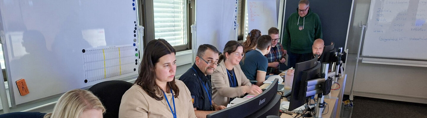Mehrere Personen sitzen in einem Büro oder Schulungsraum an einer langen Tischreihe, arbeiten an Computern und Laptops und tauschen sich teilweise miteinander aus.