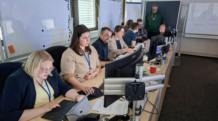Mehrere Personen sitzen in einem Büro oder Schulungsraum an einer langen Tischreihe, arbeiten an Computern und Laptops und tauschen sich teilweise miteinander aus.