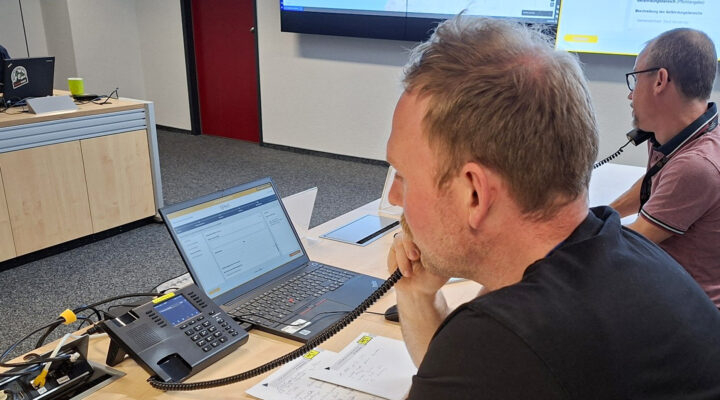 Ein Mann sitzt an einem Schreibtisch und telefoniert, während er auf einen Laptop mit geöffneter Softwareoberfläche schaut; daneben liegen Notizen und ein weiteres Telefon, im Hintergrund arbeitet eine zweite Person ebenfalls am Telefon.