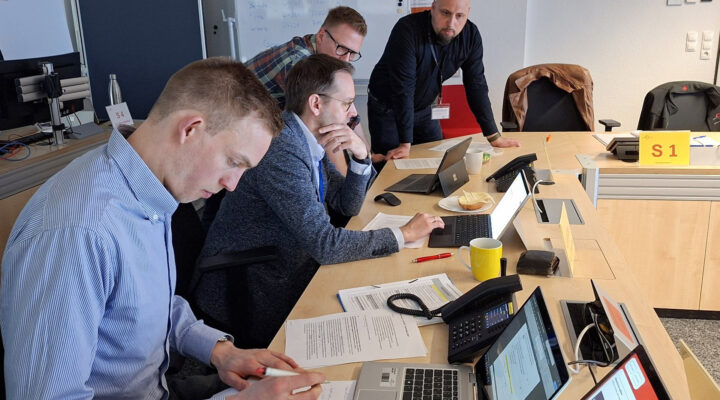 Mehrere Männer sitzen und stehen an einem Tisch in einem Büro und arbeiten gemeinsam an Laptops und Unterlagen; einer schreibt Notizen, während ein anderer sich über den Tisch beugt und auf die Bildschirme schaut.