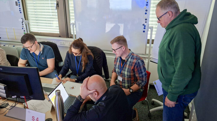 Mehrere Personen arbeiten gemeinsam an einem Tisch in einem Büro- oder Schulungsraum, nutzen Laptops und besprechen Unterlagen, während eine weitere Person daneben steht und unterstützt.