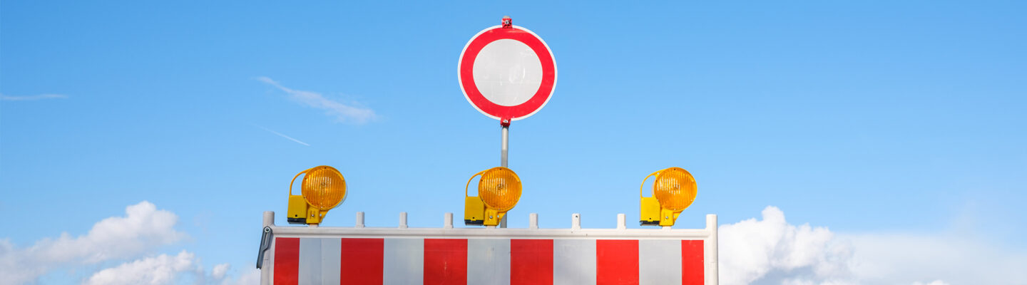 Straßensperre mit rot-weißer Absperrbake und drei gelben Warnleuchten sowie rundem Durchfahrtsverbotsschild auf einer ländlichen Straße unter blauem Himmel.