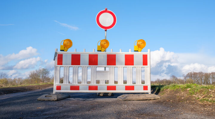 Straßensperre mit rot-weißer Absperrbake und drei gelben Warnleuchten sowie rundem Durchfahrtsverbotsschild auf einer ländlichen Straße unter blauem Himmel.