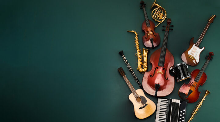 Das Bild zeigt eine dekorative Anordnung verschiedener Musikinstrumente vor einem dunkelgrünen Hintergrund. Zu sehen sind unter anderem ein Kontrabass, zwei Violinen, ein Saxophon, eine Klarinette, eine Gitarre, eine E-Gitarre, ein Akkordeon, eine Trommel, eine Trompete sowie ein Horn. Die Instrumente sind auf der rechten Bildseite gruppiert, während die linke Seite viel freie Fläche bietet.