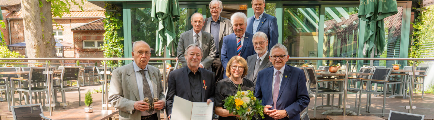 Gruppenfoto vor einem Restaurantgebäude: Mehrere ältere Männer und eine Frau stehen gemeinsam mit Landrat Olaf Meinen, während ein geehrter Mann eine Urkunde mit Verdienstorden hält und die Frau daneben einen Blumenstrauß trägt.