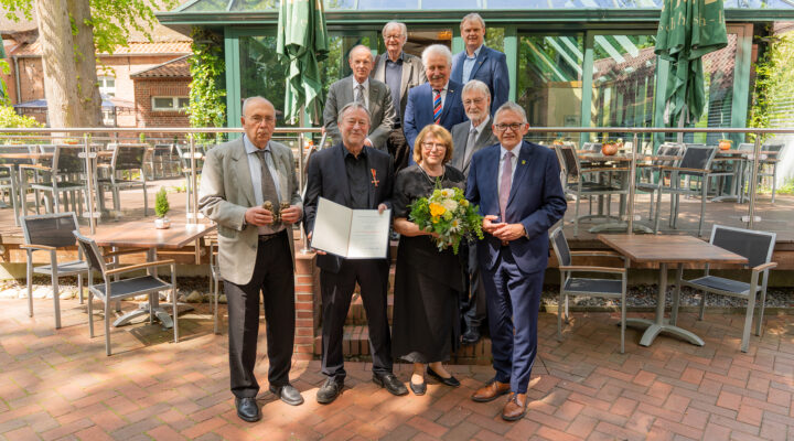 Gruppenfoto vor einem Restaurantgebäude: Mehrere ältere Männer und eine Frau stehen gemeinsam mit Landrat Olaf Meinen, während ein geehrter Mann eine Urkunde mit Verdienstorden hält und die Frau daneben einen Blumenstrauß trägt.