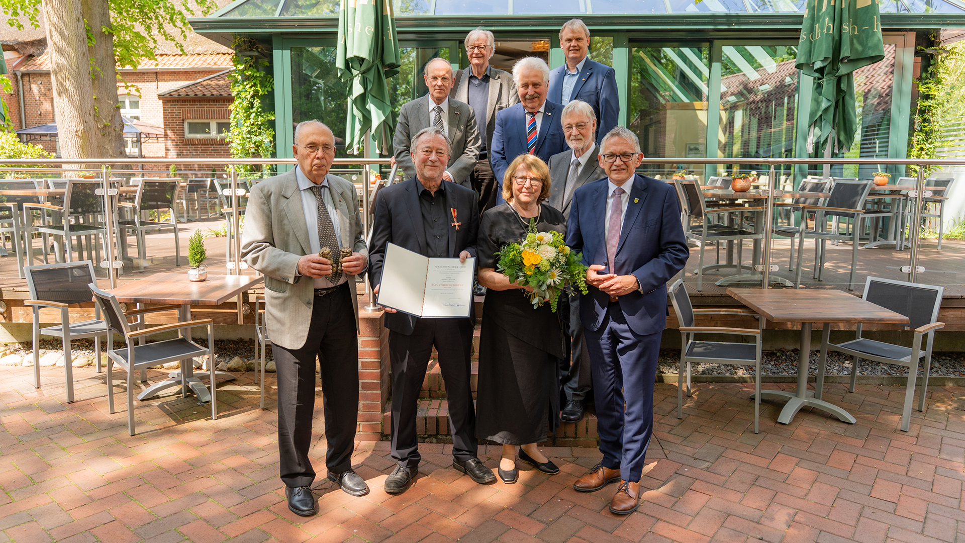 Gruppenfoto vor einem Restaurantgebäude: Mehrere ältere Männer und eine Frau stehen gemeinsam mit Landrat Olaf Meinen, während ein geehrter Mann eine Urkunde mit Verdienstorden hält und die Frau daneben einen Blumenstrauß trägt.