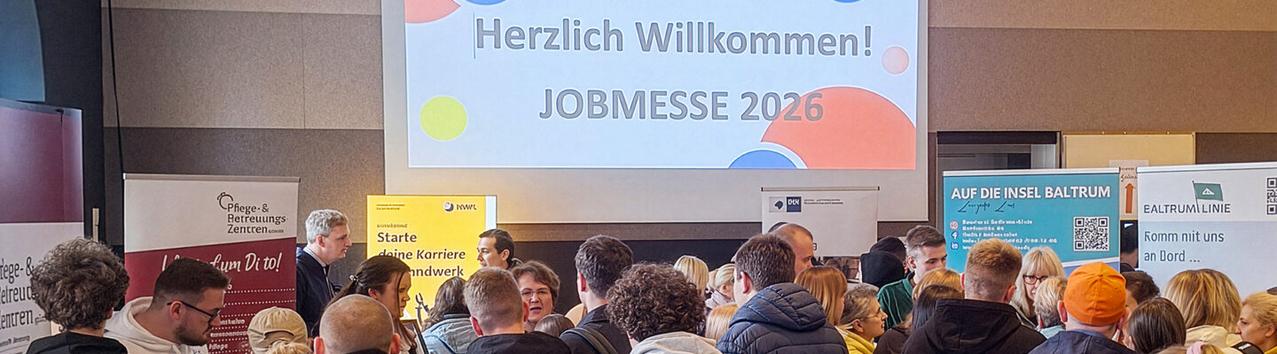 Viele Besucherinnen und Besucher stehen in einer Halle vor Informationsständen, während auf einer großen Leinwand „Herzlich Willkommen! JOBMESSE 2026“ angezeigt wird.