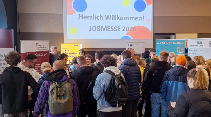 Viele Besucherinnen und Besucher stehen in einer Halle vor Informationsständen, während auf einer großen Leinwand „Herzlich Willkommen! JOBMESSE 2026“ angezeigt wird.
