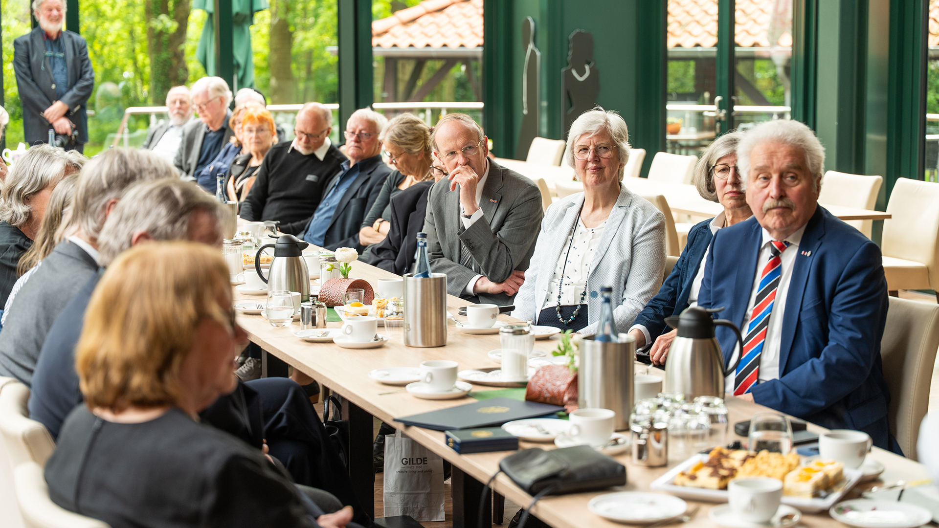 Das Foto zeigt die Gäste der Veranstaltung in einer aufmerksam lauschenden Runde an einer langen gedeckten Tafel. Im Vordergrund sitzt eine ältere Gruppe von Teilnehmerinnen und Teilnehmern, darunter ein Mann im blauen Anzug mit rot-blau gestreifter Krawatte. Die Atmosphäre wirkt ruhig, respektvoll und feierlich.