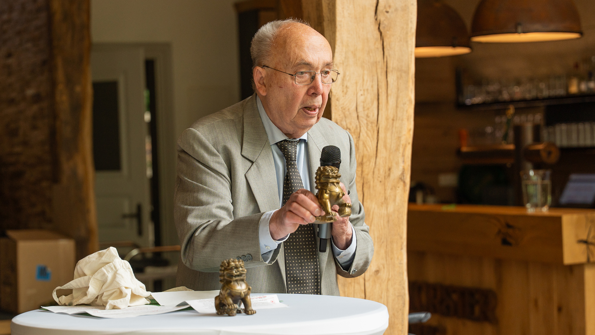 Das Bild zeigt Dr. Gerhard Stauch während seiner Ansprache bei der Veranstaltung. Er steht an einem Stehtisch mit Mikrofon und präsentiert dabei zwei kleine bronzefarbene Figuren. Die Szene wirkt persönlich und feierlich, während die warme Holzoptik des Raumes den würdevollen Charakter der Veranstaltung unterstreicht.