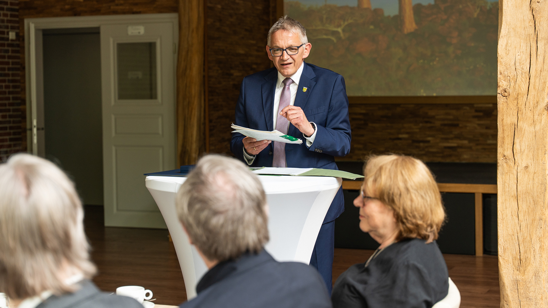 Landrat Olaf Meinen steht an einem Stehtisch und hält eine Rede vor Gästen bei einer Feierstunde. Er trägt einen blauen Anzug und hält Unterlagen in der Hand, während Zuhörerinnen und Zuhörer im Vordergrund sitzen.