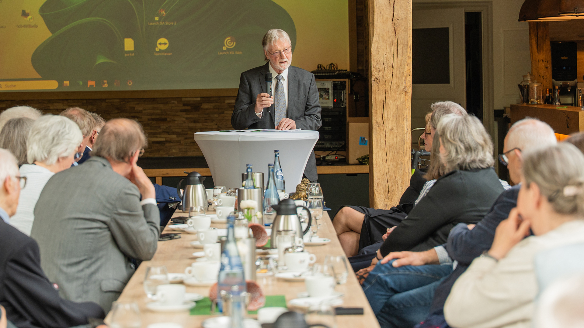 Das Bild zeigt Ulrich Kohlhoff vom Verein Gedenkstätte KZ Engerhafe e.V. während einer Rede vor einem größeren Publikum. Er steht an einem Stehtisch mit Mikrofon und Unterlagen, während die Zuhörerinnen und Zuhörer aufmerksam an langen Tafeln sitzen. Die Atmosphäre wirkt feierlich und würdevoll – passend zu einer Gedenk- oder Ehrungsveranstaltung.