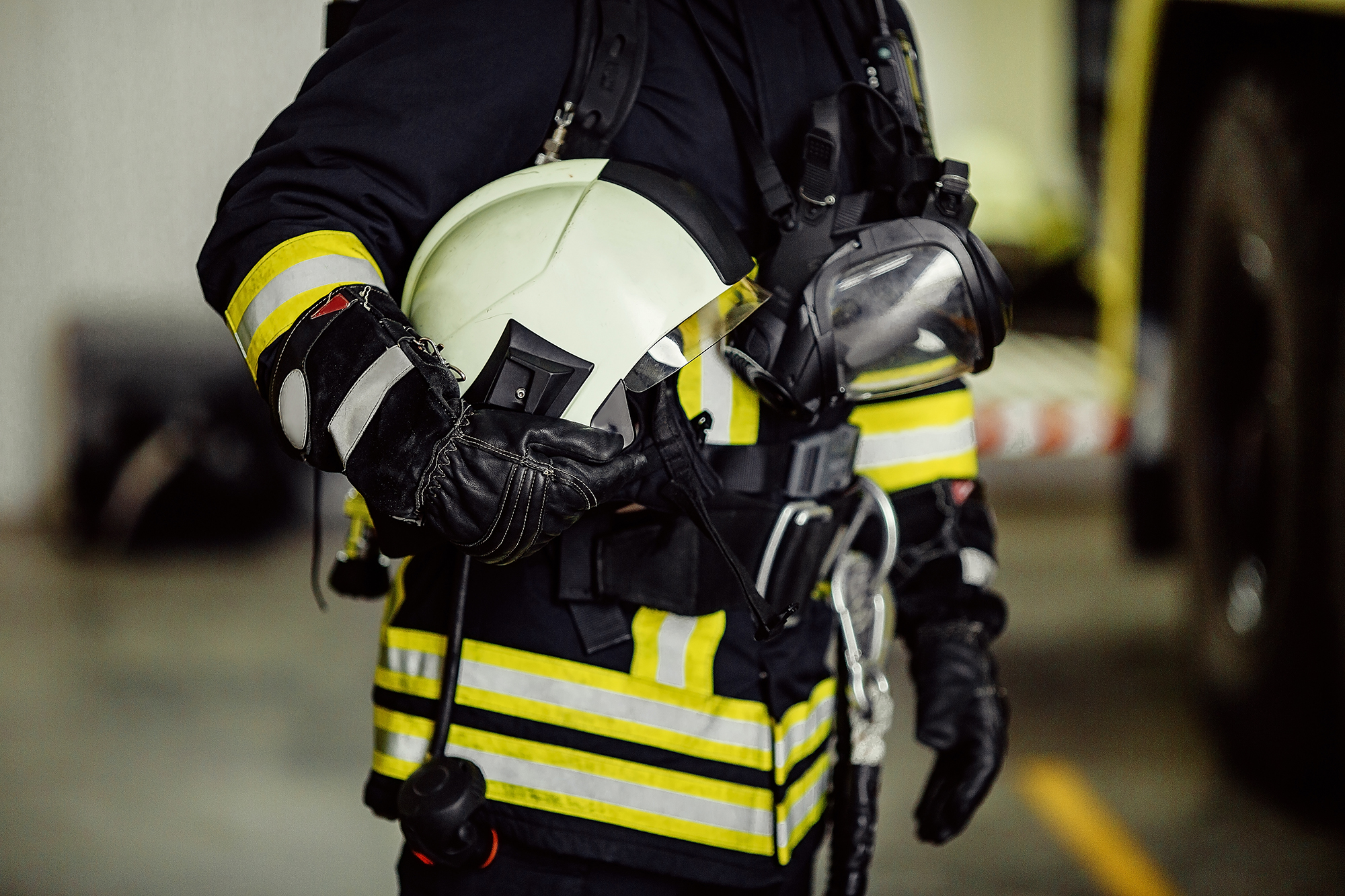 Feuerwehrkraft in Schutzkleidung mit reflektierenden Streifen hält einen weißen Helm und trägt Atemschutzgerät sowie Funkausrüstung.