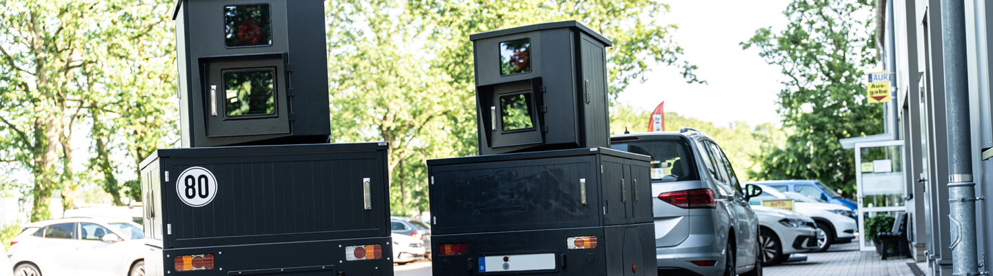 Zwei mobile Anhänger zur Geschwindigkeitsüberwachung stehen auf einem gepflasterten Parkplatz vor einem Gebäude. Die dunkelgrauen Messanlagen sind mit Kameras ausgestattet und erinnern in ihrer Form an stationäre Blitzer. Im Hintergrund sind geparkte Fahrzeuge sowie Bäume zu sehen.