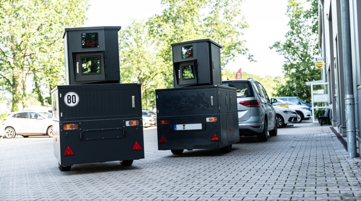 Zwei mobile Anhänger zur Geschwindigkeitsüberwachung stehen auf einem gepflasterten Parkplatz vor einem Gebäude. Die dunkelgrauen Messanlagen sind mit Kameras ausgestattet und erinnern in ihrer Form an stationäre Blitzer. Im Hintergrund sind geparkte Fahrzeuge sowie Bäume zu sehen.