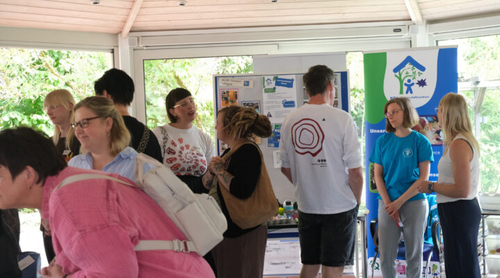 Mehrere Besucherinnen und Besucher informieren sich an verschiedenen Ständen bei einer Veranstaltung in einem hellen Veranstaltungsraum. Im Mittelpunkt stehen Gespräche zwischen den Teilnehmenden und den Ausstellenden, während Informationsbanner und Stellwände im Hintergrund verschiedene Bildungs- und Beratungsangebote präsentieren.