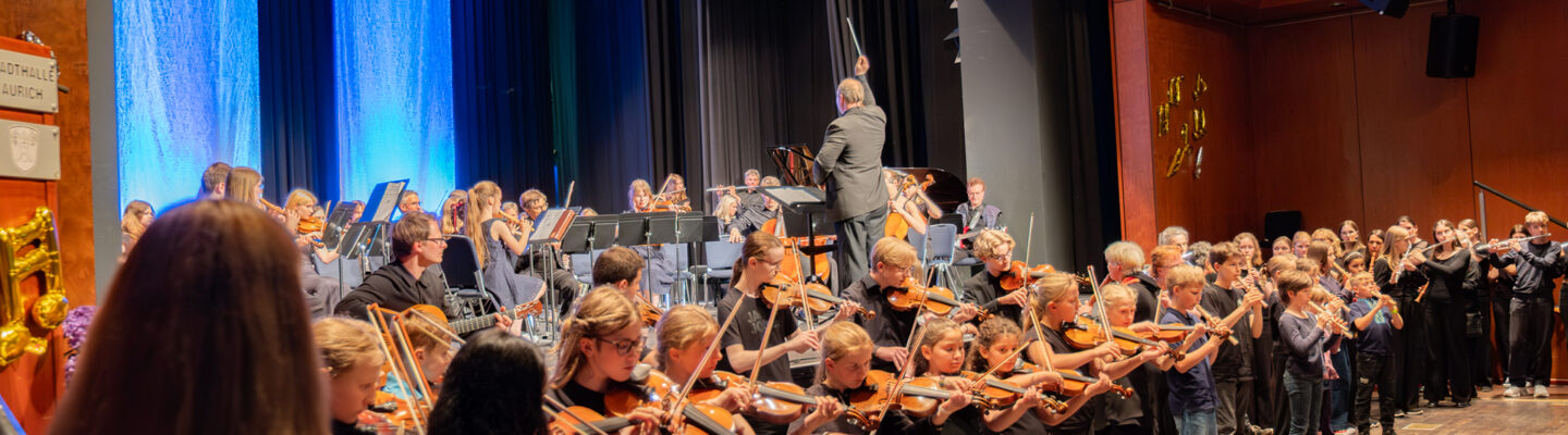 Kinder, Jugendliche und Erwachsene der Musikschule stehen und sitzen gemeinsam auf der Bühne eines Festkonzerts. Vor einem großen Orchester spielen junge Geigerinnen und Geiger, während der Dirigent das Ensemble vor blau beleuchtetem Bühnenhintergrund leitet.