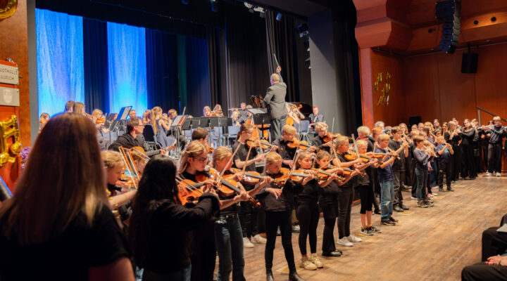 Kinder, Jugendliche und Erwachsene der Musikschule stehen und sitzen gemeinsam auf der Bühne eines Festkonzerts. Vor einem großen Orchester spielen junge Geigerinnen und Geiger, während der Dirigent das Ensemble vor blau beleuchtetem Bühnenhintergrund leitet.