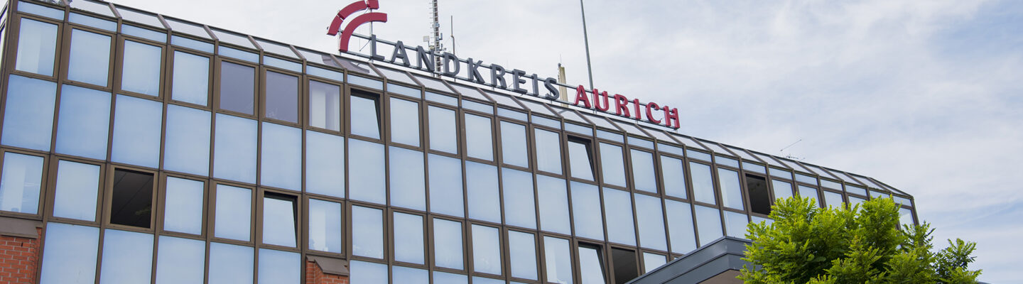 Außenansicht des Verwaltungsgebäudes des Landkreises Aurich. Auf dem Dach ist der Schriftzug „Landkreis Aurich“ angebracht, während im Vordergrund der Eingangsbereich mit Vordach und ein grüner Baum zu sehen sind. Darüber spannt sich ein leicht bewölkter Himmel.