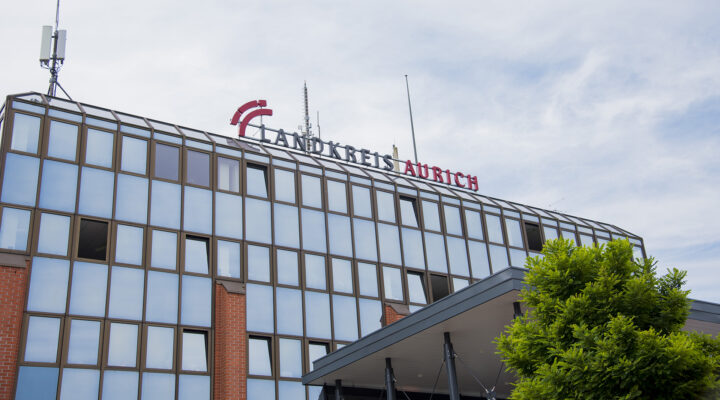 Außenansicht des Verwaltungsgebäudes des Landkreises Aurich. Auf dem Dach ist der Schriftzug „Landkreis Aurich“ angebracht, während im Vordergrund der Eingangsbereich mit Vordach und ein grüner Baum zu sehen sind. Darüber spannt sich ein leicht bewölkter Himmel.