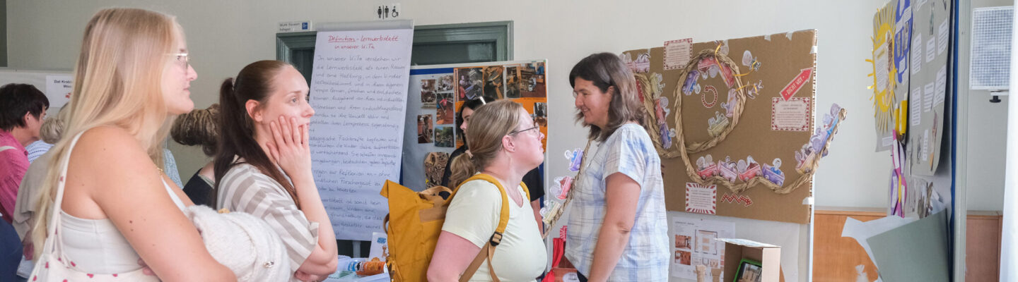 Besucherinnen und Besucher informieren sich an einem Ausstellungsstand über pädagogische Projekte und Materialien einer Kindertagesstätte. Im Gespräch mit einer Mitarbeiterin betrachten sie Präsentationstafeln, Bastelmaterialien und Beispiele aus der Lernwerkstatt.