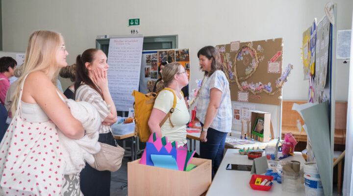 Besucherinnen und Besucher informieren sich an einem Ausstellungsstand über pädagogische Projekte und Materialien einer Kindertagesstätte. Im Gespräch mit einer Mitarbeiterin betrachten sie Präsentationstafeln, Bastelmaterialien und Beispiele aus der Lernwerkstatt.
