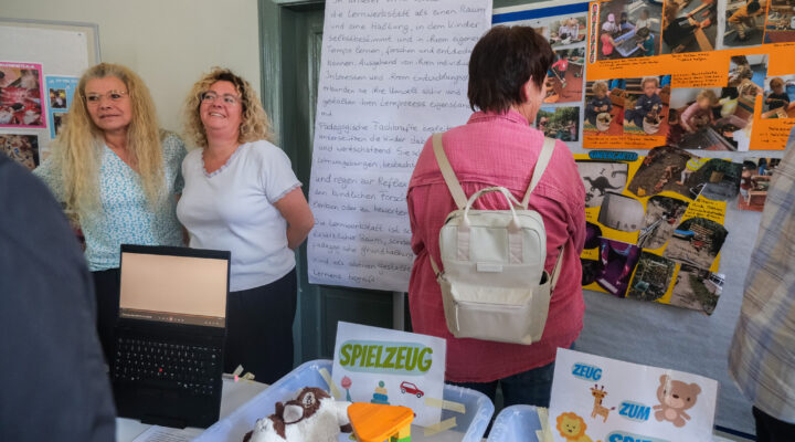 Zwei Mitarbeiterinnen einer Kindertagesstätte stehen an einem Informationsstand und erläutern Besucherinnen und Besuchern das Konzept ihrer Lernwerkstatt. Auf Stellwänden werden Fotos aus dem Kita-Alltag und pädagogische Inhalte präsentiert, während auf dem Tisch Spielmaterialien und Lernangebote ausgestellt sind.