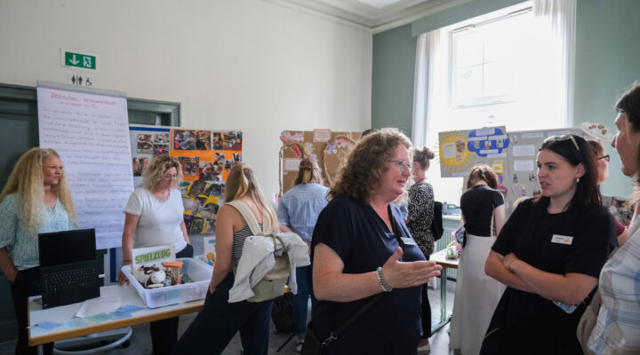 Mehrere Besucherinnen und Besucher informieren sich an verschiedenen Ständen über pädagogische Konzepte und Materialien einer Kindertagesstätte. Vor Stellwänden mit Fotos und Informationen kommen sie miteinander ins Gespräch und tauschen sich über die ausgestellten Inhalte aus.
