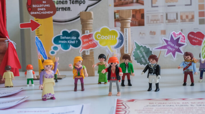 Ein Informationsstand zeigt Playmobil-Figuren mit bunten Sprechblasen, die typische Fragen und Erwartungen von Eltern und Kindern zum Schulstart darstellen. Im Hintergrund sind Infotafeln und Materialien zur Lern- und Schulentwicklung zu sehen.