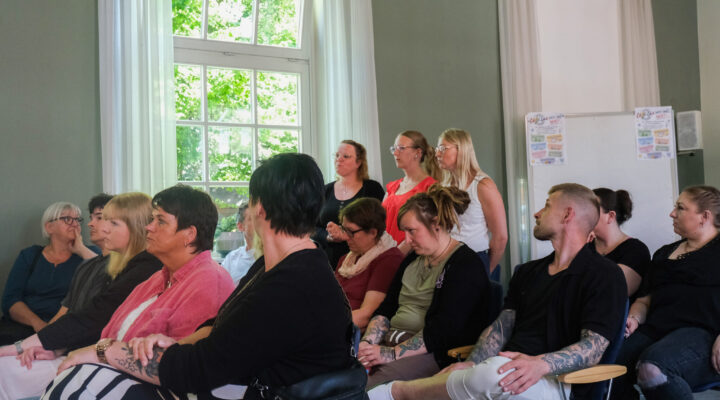 Mehrere Teilnehmerinnen und Teilnehmer sitzen und stehen in einem hellen Veranstaltungsraum und verfolgen aufmerksam einen Vortrag. Einige hören konzentriert zu, während drei Frauen im Hintergrund stehen und das Geschehen beobachten. Große Fenster sorgen für viel Tageslicht.