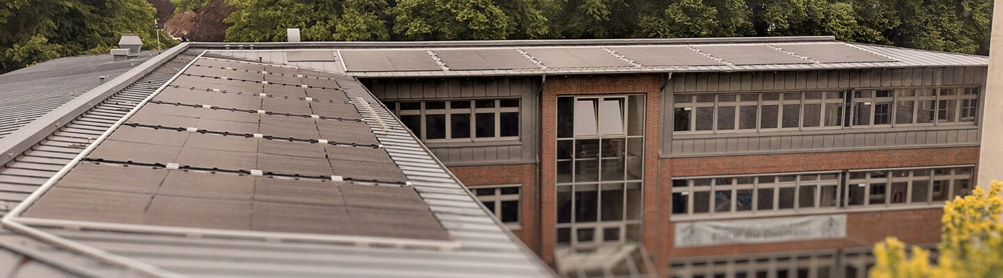 Auf dem Dach eines Schulgebäudes sind mehrere Reihen von Photovoltaikmodulen installiert. Die Aufnahme zeigt die Solaranlage aus erhöhter Perspektive, während im Hintergrund das Schulgebäude und große Bäume zu sehen sind.