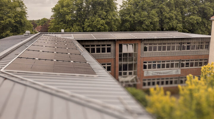 Auf dem Dach eines Schulgebäudes sind mehrere Reihen von Photovoltaikmodulen installiert. Die Aufnahme zeigt die Solaranlage aus erhöhter Perspektive, während im Hintergrund das Schulgebäude und große Bäume zu sehen sind.