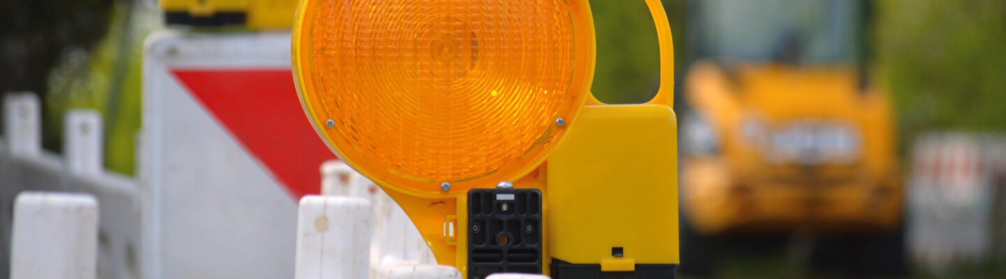 Nahaufnahme einer gelben Warnleuchte an einer rot-weiß markierten Baustellenabsperrung. Im unscharfen Hintergrund ist eine Straßenbaustelle mit einer Baumaschine zu erkennen.