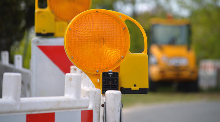 Nahaufnahme einer gelben Warnleuchte an einer rot-weiß markierten Baustellenabsperrung. Im unscharfen Hintergrund ist eine Straßenbaustelle mit einer Baumaschine zu erkennen.