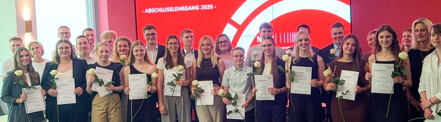 Gruppenfoto der Absolventinnen und Absolventen des Abschlusslehrgangs 2026 für Verwaltungsfachangestellte am Lehrgangsort Aurich. Die Teilnehmenden stehen gemeinsam mit Lehrkräften und Begleitpersonen vor einer großen roten Präsentationswand. Viele halten ihre Abschlusszeugnisse und weiße Rosen in den Händen und blicken lächelnd in die Kamera. Auf dem Bildschirm im Hintergrund steht: „Unsere Verwaltungsfachangestellten vom Lehrgangsort Aurich – Abschlusslehrgang 2026“.