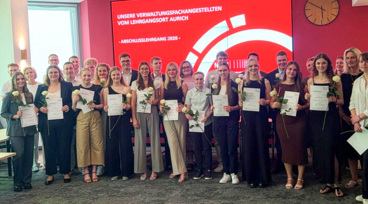 Gruppenfoto der Absolventinnen und Absolventen des Abschlusslehrgangs 2026 für Verwaltungsfachangestellte am Lehrgangsort Aurich. Die Teilnehmenden stehen gemeinsam mit Lehrkräften und Begleitpersonen vor einer großen roten Präsentationswand. Viele halten ihre Abschlusszeugnisse und weiße Rosen in den Händen und blicken lächelnd in die Kamera. Auf dem Bildschirm im Hintergrund steht: „Unsere Verwaltungsfachangestellten vom Lehrgangsort Aurich – Abschlusslehrgang 2026“.