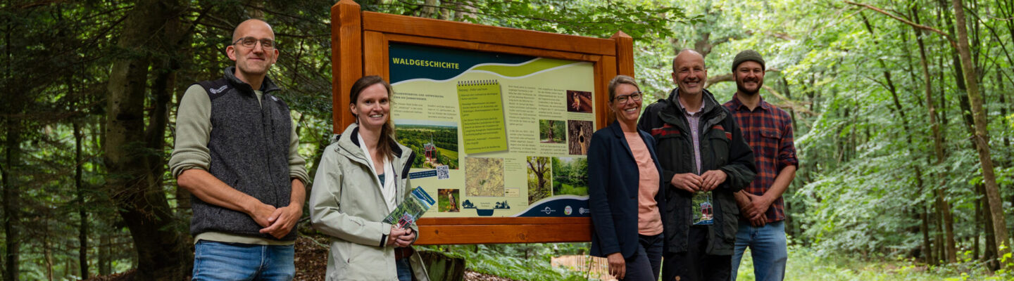 Fünf Personen stehen auf einem Waldweg neben einer großen Informationstafel mit der Überschrift „Waldgeschichte“. Sie lächeln in die Kamera und halten teilweise Informationsflyer in den Händen. Die Tafel ist in einen hölzernen Rahmen eingefasst und vermittelt Wissenswertes zur Entwicklung des Waldes. Umgeben von dichtem Mischwald mit hohen Bäumen und grünem Blätterdach vermittelt die Szene den Eindruck einer gemeinsamen Vorstellung oder Eröffnung eines Naturinformationsangebots.
