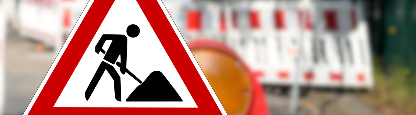 Nahaufnahme eines dreieckigen Verkehrszeichens „Baustelle“ mit rotem Rand und schwarzem Piktogramm eines Bauarbeiters. Im unscharfen Hintergrund sind rot-weiße Absperrbaken und weitere Baustellenabsicherungen entlang einer Straße zu erkennen. Das Warnschild steht im Vordergrund und weist deutlich auf Straßenbauarbeiten hin.