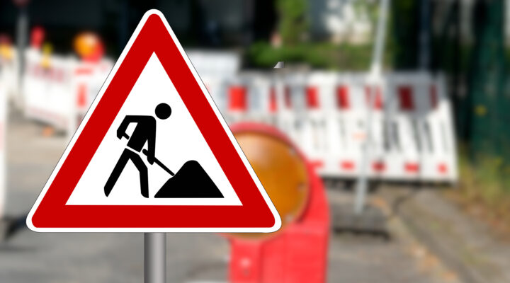 Nahaufnahme eines dreieckigen Verkehrszeichens „Baustelle“ mit rotem Rand und schwarzem Piktogramm eines Bauarbeiters. Im unscharfen Hintergrund sind rot-weiße Absperrbaken und weitere Baustellenabsicherungen entlang einer Straße zu erkennen. Das Warnschild steht im Vordergrund und weist deutlich auf Straßenbauarbeiten hin.