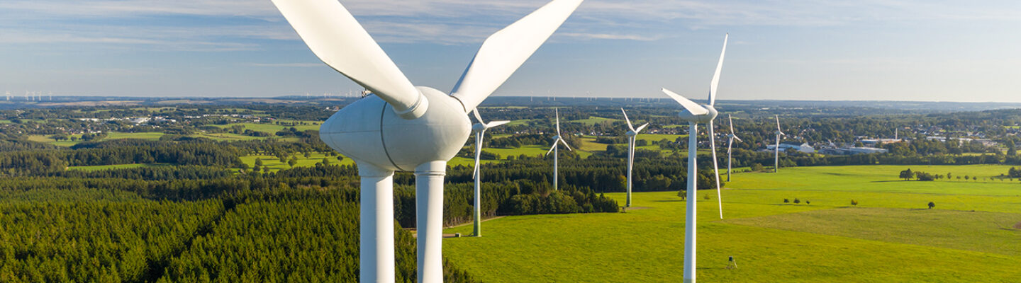 Luftaufnahme eines Windparks mit mehreren modernen Windenergieanlagen in einer weitläufigen, grünen Landschaft. Im Vordergrund dominiert eine Windkraftanlage mit ihren weißen Rotorblättern das Bild. Dahinter stehen weitere Anlagen zwischen Wiesen, Feldern und einem Waldstück. Am Horizont sind verstreute Ortschaften und zusätzliche Windräder unter einem klaren, blau-weißen Himmel zu erkennen.