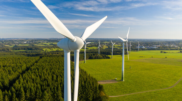 Luftaufnahme eines Windparks mit mehreren modernen Windenergieanlagen in einer weitläufigen, grünen Landschaft. Im Vordergrund dominiert eine Windkraftanlage mit ihren weißen Rotorblättern das Bild. Dahinter stehen weitere Anlagen zwischen Wiesen, Feldern und einem Waldstück. Am Horizont sind verstreute Ortschaften und zusätzliche Windräder unter einem klaren, blau-weißen Himmel zu erkennen.