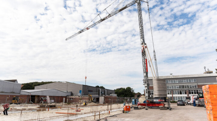 Großbaustelle mit einem hohen Turmdrehkran unter bewölktem Himmel. Auf dem Gelände sind bereits Fundamente und Stahlbewehrungen angelegt; im Hintergrund stehen Gewerbegebäude, Baumaschinen und weitere Baustelleneinrichtungen.